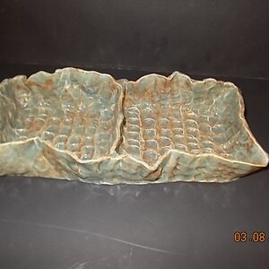 May include: A ceramic serving dish with two sections. The dish is a light blue-green color with a textured surface. The dish has a unique, organic shape.