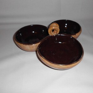 May include: A set of three brown ceramic bowls with a unique design. The bowls are arranged in a clover shape, with a handle connecting them. The interior of the bowls is a dark brown, contrasting with the lighter exterior.