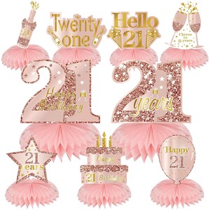 May include: A collection of rose gold glittery 21st birthday decorations. Includes a champagne bottle, number cutouts, a star, a cake, and a balloon, all with the number 21. Each piece sits atop a pink honeycomb base.