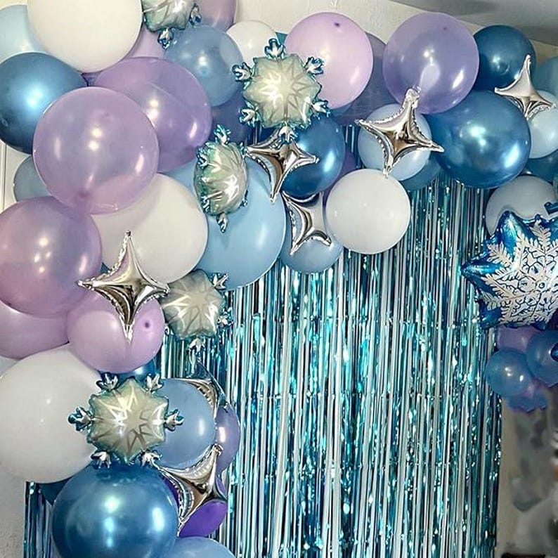 Puede incluir: Un arco de globos festivo en tonos azules, morados y blancos, con detalles de estrellas y copos de nieve plateados. Los globos est&aacute;n dispuestos en cascada, creando un ambiente festivo. Una cortina azul brillante sirve de fondo.