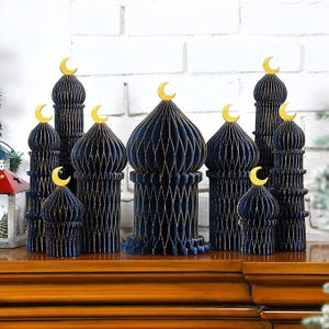 May include: A collection of navy blue paper mosque decorations with gold accents and crescent moon toppers. The decorative pieces are arranged on a wooden surface in front of a white brick wall. The mosque decorations vary in size and shape.