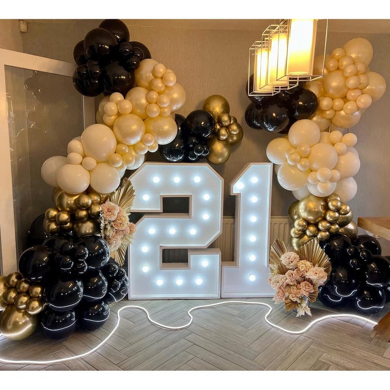 May include: A celebratory display featuring a large, illuminated number "21" with surrounding balloon arches in black, gold, and cream colors. Floral accents and a glowing light cord add to the festive atmosphere, perfect for a birthday celebration.