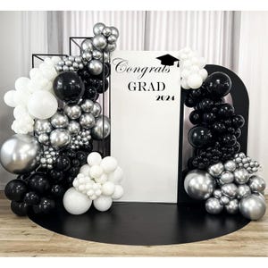 May include: A graduation party backdrop with black, white, and silver balloons. The backdrop features a white sign with the words "Congrats GRAD 2024" and a graduation cap graphic. The balloons are arranged in clusters and arches.