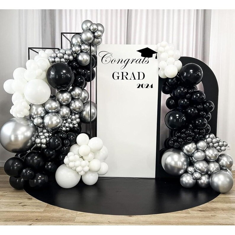 May include: A graduation party backdrop with black, white, and silver balloons. The backdrop features a white sign with the words "Congrats GRAD 2024" and a graduation cap graphic. The balloons are arranged in clusters and arches.
