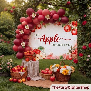 May include: A rectangular sign with the text THE Apple OF OUR EYE, decorated with red and burgundy balloons, white flowers, and apple illustrations. Below are two woven baskets filled with red apples and a basket of yellow and white flowers.