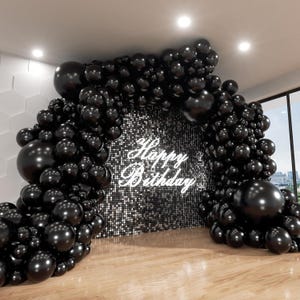 May include: A black balloon arch frames a backdrop with the words "Happy Birthday" in white script. The backdrop is covered in small, reflective squares. The arch is set on a wooden floor.