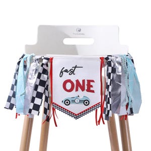 May include: A high chair banner with a racing theme. The banner is white with the words "fast ONE" in black and red. A blue race car is below the text. The banner is decorated with checkered flags, silver streamers, and blue fabric.