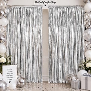 May include: Two metallic silver foil fringe curtains hanging side by side, flanked by clusters of silver and white balloons. A white sign with black text reads: Good Friends Brighter Birthdays Happier Times. Two gift boxes sit on the floor.