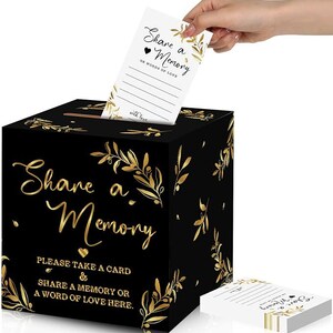 May include: Black box with gold floral design and the text "Share a Memory" on the front. The box has a slot for inserting cards. A white card with the text "Share a Memory" and "Or words of love" is being inserted into the box.