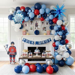 May include: A festive hockey-themed party setup with a balloon arch in blue, red, and silver. A banner reads "Puck Drop" above a table with treats and a cake. A cutout of a hockey player stands nearby.