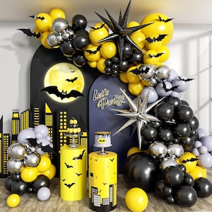 May include: A Batman-themed party decoration set with yellow, black, and silver balloons, bat silhouettes, and a cityscape backdrop. Includes a cake and decorative stands. The words "Let's Party!" are visible.