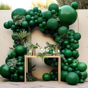 May include: A green balloon arch with greenery and a wooden table in front of a beige wall. The balloons are arranged in a cascading pattern, creating a festive backdrop.