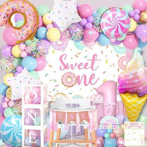 May include: A pink and white 'Sweet One' banner with donut and sprinkle decorations. A clear box with the word 'ONE' in white letters is on a table with a pink and white 'ONE' banner. There are pink, white, and blue balloons with donut and sprinkle designs.