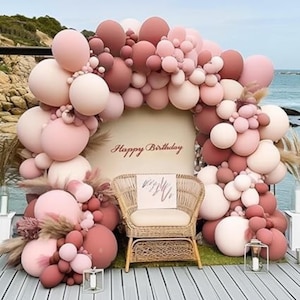 May include: A festive birthday scene with a large balloon arch in shades of pink and mauve. A woven chair sits in front of a backdrop that reads "Happy Birthday". Decorative candles and pampas grass add to the celebratory atmosphere.