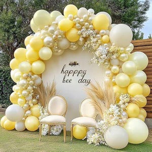 May include: A festive outdoor scene with a balloon arch in shades of yellow and cream, framing a backdrop that reads "happy bee day." Two elegant chairs are positioned in front, complemented by floral arrangements and pampas grass. The overall theme suggests a celebration.