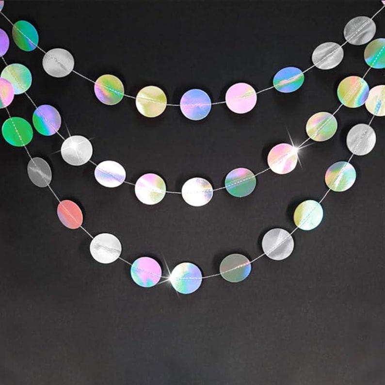 May include: Three strands of iridescent, circular garland hang against a black background. The discs reflect a rainbow of colors, including pink, green, and silver. The garland is strung on thin, clear cords, creating a festive and decorative effect.