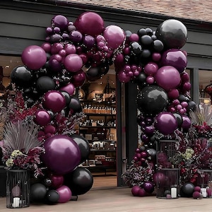 May include: A decorative archway made of black and deep purple balloons, with accents of dark red foliage. The balloons are arranged in a cascading design, creating a festive and elegant entrance. The archway is set against a dark background.