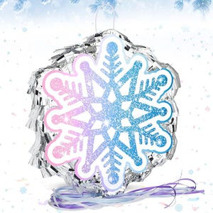 May include: A snowflake-shaped piñata with a gradient of pink and blue glitter. The snowflake is outlined in white and surrounded by silver fringe. Purple and white ribbons hang from the bottom, and a white string is at the top for hanging.