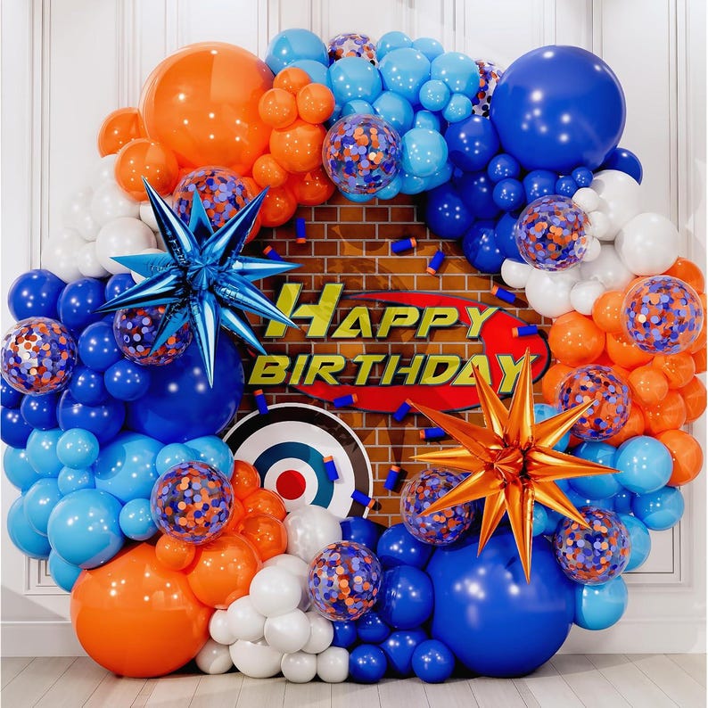 May include: A festive birthday balloon arch in shades of blue, orange, and white. The arrangement includes star-shaped decorations and confetti-filled balloons. The words "HAPPY BIRTHDAY" are displayed on a brick-patterned backdrop.