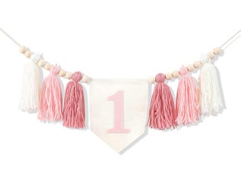 May include: A banner with a white pennant featuring a pink number 1. The banner is decorated with pink, white, and light pink tassels and wooden beads, perfect for a first birthday celebration.
