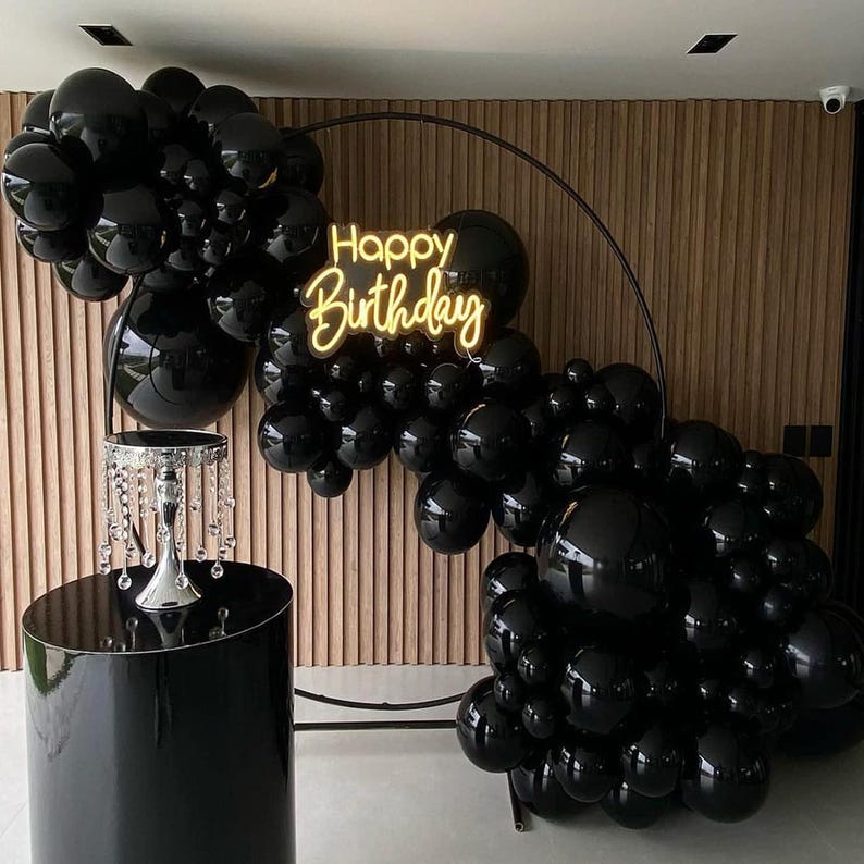 May include: A black balloon arch with a neon "Happy Birthday" sign. The arch is set against a wooden panel backdrop, with a black pedestal table holding a decorative crystal piece. The balloons are clustered in various sizes.