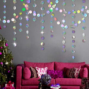 May include: Iridescent, circular garland strands hang against a gray wall, reflecting light. A magenta sofa with decorative pillows sits below. A Christmas tree with purple ornaments is on the left.