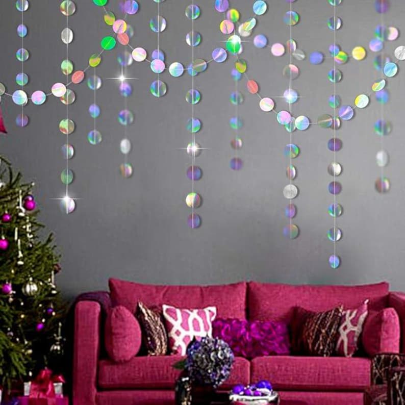 May include: Iridescent, circular garland strands hang against a gray wall, reflecting light. A magenta sofa with decorative pillows sits below. A Christmas tree with purple ornaments is on the left.
