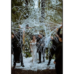 May include: A wedding ceremony with a bride in a white gown and a groom surrounded by guests. White streamers are being released over the couple. The setting is outdoors, with trees and snow on the ground.