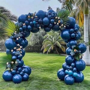 May include: A navy blue balloon arch decorated with various sizes of balloons, greenery, and small floral accents. The arch is set up outdoors on a grassy lawn, with palm trees in the background. The balloons are arranged in an arch shape.