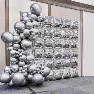 May include: A silver balloon arch and backdrop. The arch is made of various sized round balloons. The backdrop is a square of silver, quilted-looking material. The balloons and backdrop are set up on a gray surface.