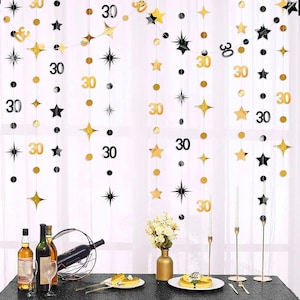 May include: Gold, black, and silver star and circle garland with the number 30. The decorations are hanging in front of a window. A table is set with wine bottles, glasses, and a cake.