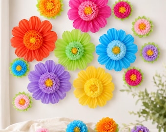 May include: A collection of colorful paper flowers in various sizes and hues, including orange, pink, blue, green, yellow, and purple. The flowers are arranged on a white wall and a white surface, creating a vibrant and cheerful display.