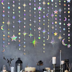 May include: Holographic star and moon garland hanging against a dark gray wall. The garland features various star shapes, crescent moons, and circular accents in iridescent colors. Decorative candles and a lantern are visible below.