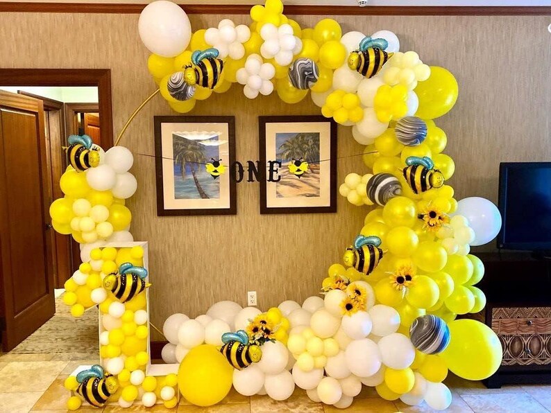 Puede incluir: Decoraci&oacute;n festiva para fiestas con una gran disposici&oacute;n de globos amarillos y blancos, con detalles de abejas y flores. La exhibici&oacute;n incluye un gran n&uacute;mero "1" y un arco circular, con la palabra "ONE" en una pancarta.