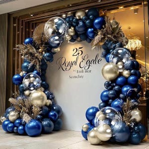 May include: An arch-shaped balloon arrangement in shades of blue and gold, with clear bubble balloons and dried floral accents. The arrangement features the text "25 Royal Egale" in elegant script, suitable for a celebration or event.