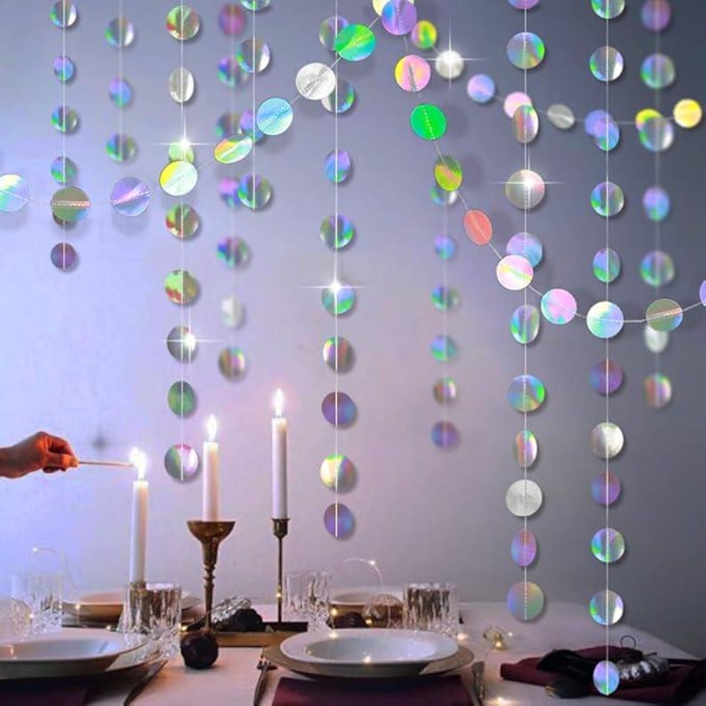 May include: Iridescent circle garlands hanging as a backdrop for a table setting. White candles in glass holders and a person lighting a match are visible. The table is set with plates and silverware.