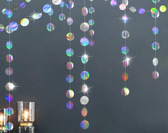 May include: Iridescent, circular garland strands hang against a dark gray wall. The decorative strands feature a rainbow of colors and reflect light. The garland is a festive decoration, perfect for parties and celebrations.