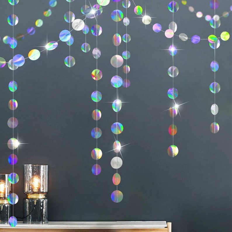 May include: Iridescent, circular garland strands hang against a dark gray wall. The decorative strands feature a rainbow of colors and reflect light. The garland is a festive decoration, perfect for parties and celebrations.