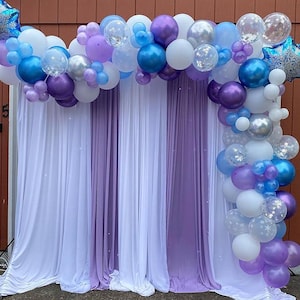 Puede incluir: Decoraci&oacute;n de fiesta con un arco de globos en tonos azules, morados y blancos. Los globos est&aacute;n dispuestos sobre un tel&oacute;n de fondo de cortinas de tela blancas y moradas, creando un ambiente festivo.