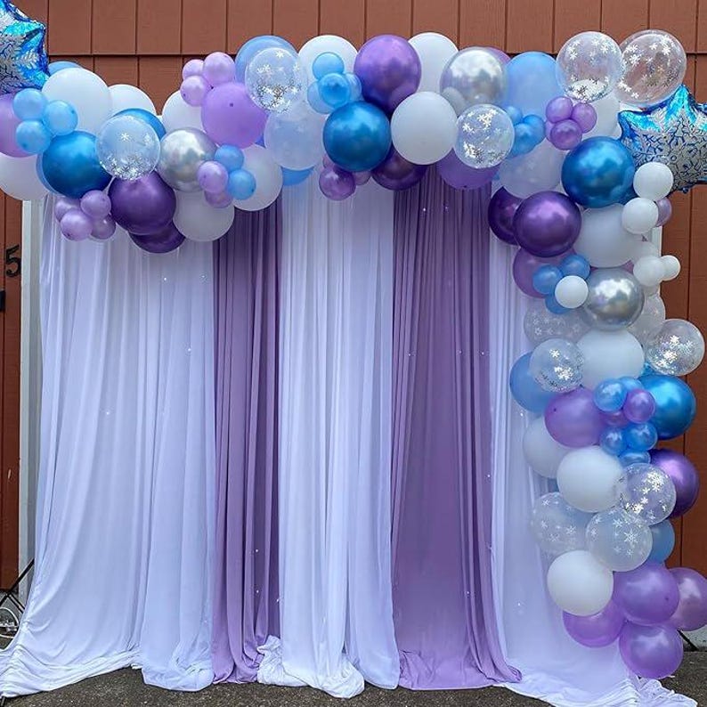 Puede incluir: Decoraci&oacute;n de fiesta con un arco de globos en tonos azules, morados y blancos. Los globos est&aacute;n dispuestos sobre un tel&oacute;n de fondo de cortinas de tela blancas y moradas, creando un ambiente festivo.