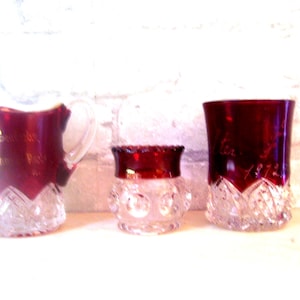 May include: Three vintage glass pieces with a ruby red color and clear glass bases. The pieces are a pitcher, a small bowl, and a tumbler. The pitcher and tumbler have gold lettering on the side.