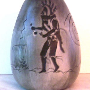 May include: A gray, egg-shaped ceramic vase with a dark silhouette of a Native American figure holding objects. The vase has etched designs of a spiral, a zigzag, and a plant. The vase has a smooth, glossy finish.