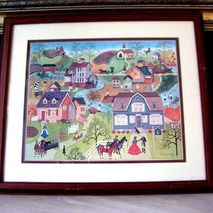 May include: A framed print of a colorful folk art painting depicting a quaint village scene with houses, a church, a bridge, a river, trees, and people. The painting is in a red wooden frame.