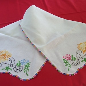 May include: White embroidered handkerchief with colorful floral designs in pink, yellow, and blue. The edges are scalloped and trimmed with a rainbow of colors. The handkerchief is folded on a red surface.