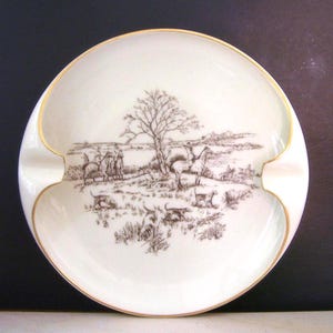 May include: A white ceramic ashtray with a gold rim and a brown and white illustration of a hunting scene with horses and dogs.