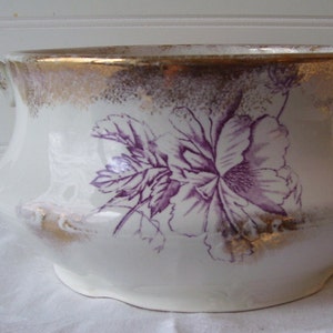May include: A white ceramic chamber pot with a purple floral design and gold accents. The pot has a handle and a rounded shape.