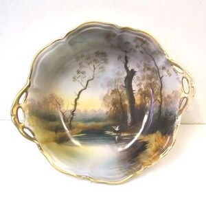 May include: A decorative, scalloped-edge porcelain dish with a hand-painted landscape scene. The artwork depicts a serene waterside view with trees and a soft, golden-yellow sky. The rim is trimmed with gold.