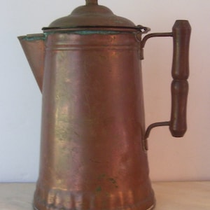May include: A copper coffee pot with a wooden handle and a lid. The pot has a slightly tarnished finish and a fluted base.