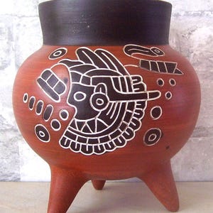 May include: A terracotta pot with three legs and a black rim. The pot is decorated with a black and white design of a stylized bird or animal with geometric patterns.