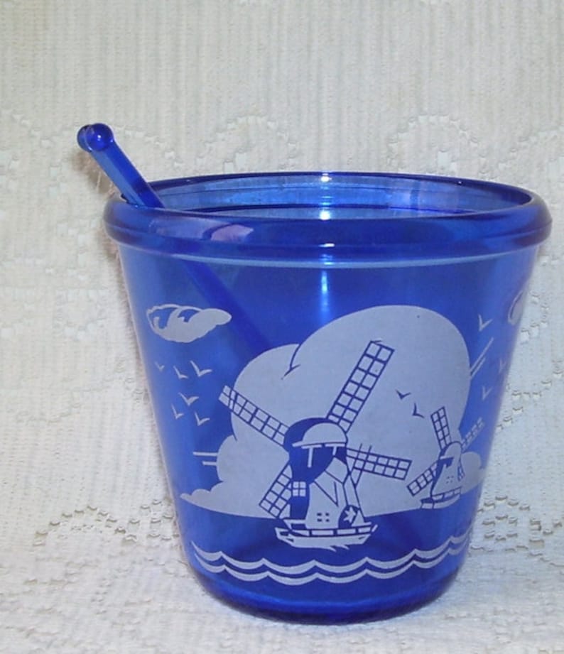 May include: A cobalt blue glass cup with a white windmill design and a matching blue plastic stirrer. The cup has a white rim and a white base.